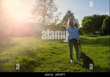 Une femme vêtue de blanc marche son petit chien noir dans un parc de Londres au crépuscule dans l'été Banque D'Images