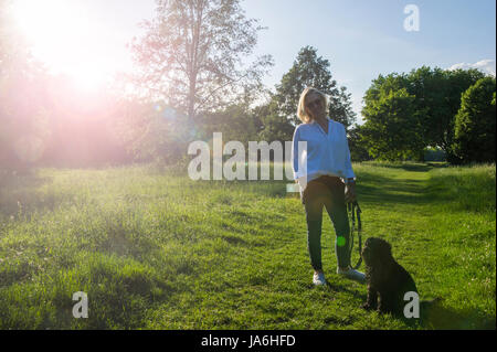 Une femme vêtue de blanc marche son petit chien noir dans un parc de Londres au crépuscule dans l'été Banque D'Images