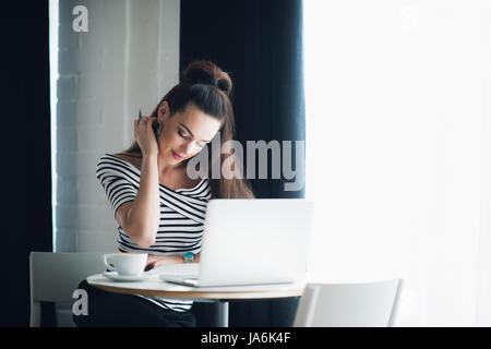 Tourné à l'intérieur du charmant jeune femme avec des cheveux chocolat watch portant sur son poignet, assis à un café et à l'aide de l'ordinateur portable générique, lumière du jour fenêtre creux en arrière-plan. Banque D'Images