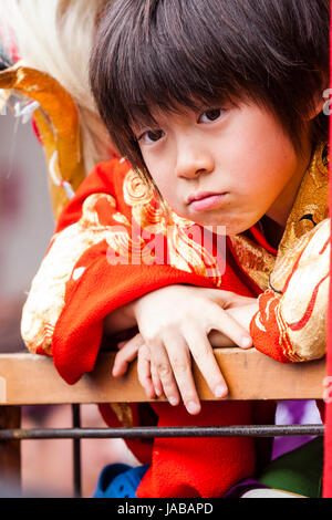 L'enfant japonais, garçon, 8-9 ans, penché sur la rampe en bois à directement au spectateur, malheureux, en colère, l'expression dans les yeux. Yukata rouge porte manteau. Banque D'Images