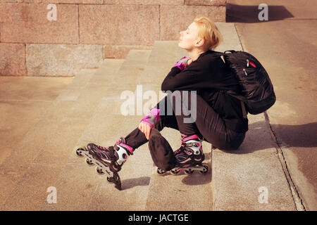 Jeune fille en noir casual avec patins à assis sur l'escalier. L'extérieur sunny shoot Banque D'Images