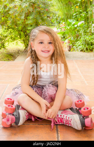 École maternelle Petite fille assise sur sol portant des patins à son passage et ses jambes, dans un fond de jardin Banque D'Images