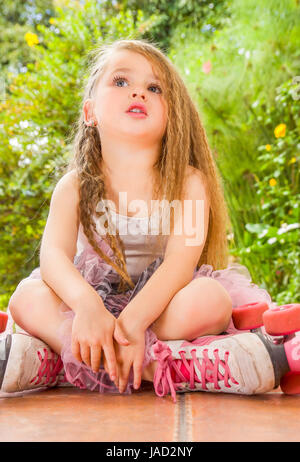 Petite fille assise sur le sol portant des patins à son passage et ses jambes à la recherche le ciel, dans un fond de jardin Banque D'Images