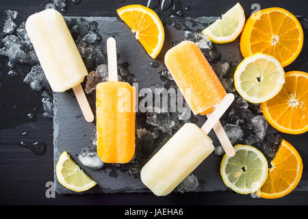 Orange et citron sucettes faites maison avec de la glace et d'agrumes sur fond noir en pierre. Concept des aliments en été. Vue d'en haut. Banque D'Images