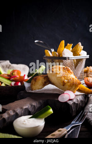 Fish and chips. Filet de poisson frit avec des pommes de terre au four et une salade fraîche sur planche à découper en bois. Banque D'Images