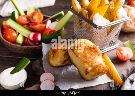 Fish and chips. Filet de poisson frit avec des pommes de terre au four et une salade fraîche sur planche à découper en bois. Banque D'Images