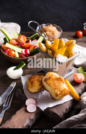 Fish and chips. Filet de poisson frit avec des pommes de terre au four et une salade fraîche sur planche à découper en bois. Banque D'Images