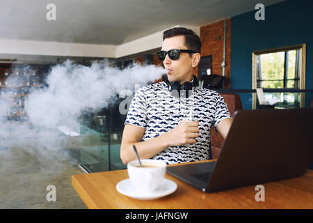 Beau jeune homme hipster avec barbe, assis dans un café avec une tasse de café, seine et libère un nuage d'vaporsitting. Au travail et d'avoir l'ordinateur portable Banque D'Images