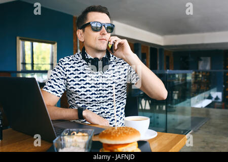 Beau jeune homme hipster dans sunglases assis dans cafe parle téléphone mobile, ordinateur portable sur une table en bois. Banque D'Images