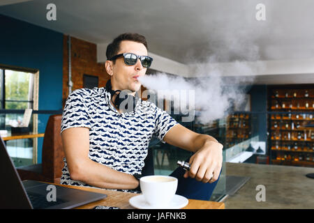 Beau jeune homme hipster avec barbe, assis dans un café avec une tasse de café, seine et libère un nuage d'vaporsitting. Au travail et d'avoir l'ordinateur portable Banque D'Images