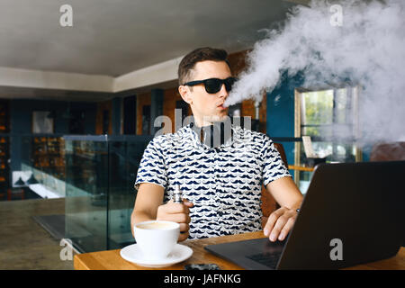 Beau jeune homme hipster avec barbe, assis dans un café avec une tasse de café, seine et libère un nuage d'vaporsitting. Au travail et d'avoir l'ordinateur portable Banque D'Images