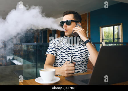 Beau jeune homme hipster avec barbe, assis dans un café avec une tasse de café, seine et libère un nuage d'vaporsitting. Au travail et d'avoir l'ordinateur portable Banque D'Images