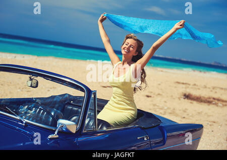 Femme heureuse avec un mains levées assis en voiture cabriolet rétro sur la plage près de la mer turquoise. Paysage idyllique Banque D'Images