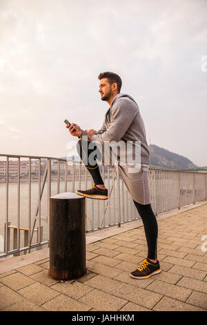 Un beau jeune homme mettant sa jambe vers le haut et se penchant sur elle tout en utilisant son téléphone du riverside Banque D'Images