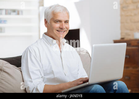 Smiling mature homme assis sur un canapé de la saisie sur un ordinateur portable dans une salle de séjour Banque D'Images