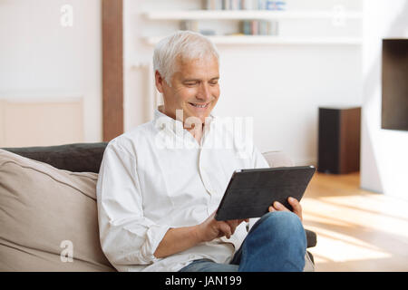 Smiling mature homme assis sur un canapé à l'aide d'un tablet pc dans un salon Banque D'Images