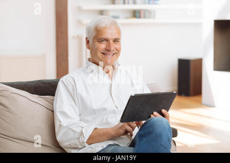 Smiling mature homme assis sur un canapé à l'aide d'un tablet pc dans un salon Banque D'Images