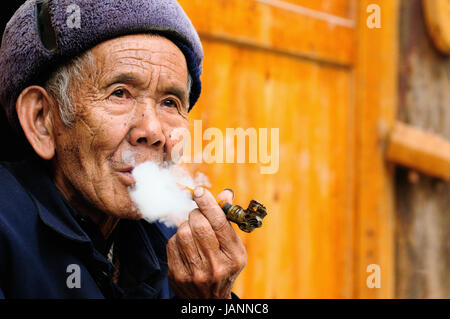 ZHAOXING, CHINE - 11 NOVEMBRE 2010 : Old Chinese reposant sur la porte de son chalet fumant une pipe dans le village Zhaoxing Banque D'Images