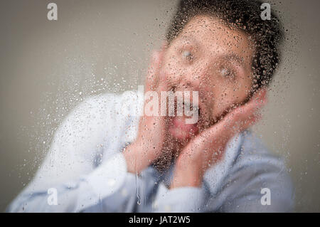 Jeune homme crier à l'aide de ses deux mains sur son visage, derrière une vitre brouillée avec des gouttes, fond gris. Banque D'Images