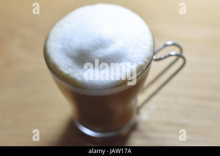 Crème mousseuse au sommet d'un cappuchino dans une tasse en verre italien avec poignée en acier inoxydable. Banque D'Images