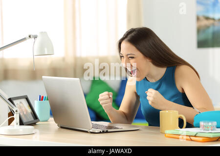 Seul élève excité à la ligne avec un ordinateur portable assis dans un bureau dans sa chambre dans une maison intérieur Banque D'Images