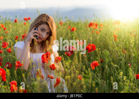 Young Girl wearing white dress cloth debout dans un champs de coquelicots avec son téléphone Banque D'Images