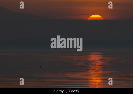 Magnifique coucher de soleil avec la moitié du soleil descendant derrière certaines montagnes et réfléchir sur un lac, avec des tons rouge et orange dans le ciel et nuages Banque D'Images