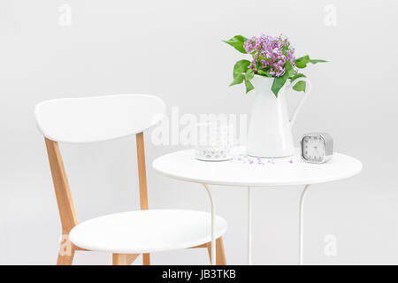 L'intérieur élégant avec table et chaise, blanc décoré d'un bouquet de lilas mauve, bougie et réveil. Banque D'Images