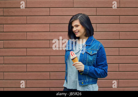 Jeune fille mignonne tenant un cornet de crème glacée avec de la confiture à la main. Femme sur un mur de briques de base sur la rue d'un denim jacket Banque D'Images