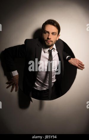 Beau jeune homme barbu randonnées hors d'un trou circulaire dans un mur blanc dans une image conceptuelle Banque D'Images