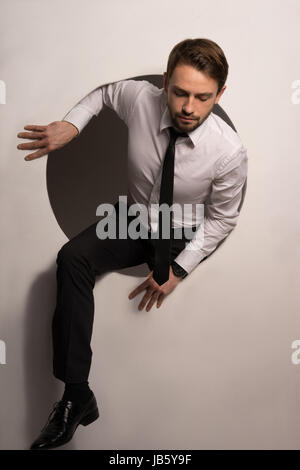 Beau jeune homme barbu randonnées hors d'un trou circulaire dans un mur blanc dans une image conceptuelle Banque D'Images