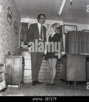 Années 1970, historique, d'un secrétaire portant une mini-jupe et avec un buoffant la coiffure se tient à côté de son grand homme patron dans son bureau. Banque D'Images