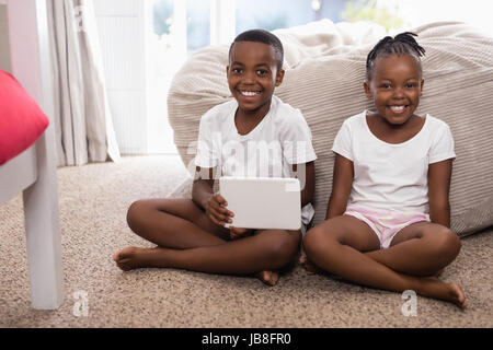 Portrait of happy siblings with digital tablet assis sur un tapis à la maison Banque D'Images