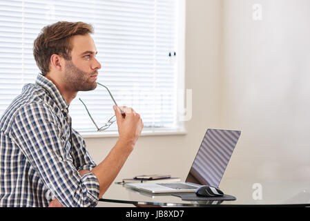 Beau jeune homme assis derrière son ordinateur portable, à la recherche dans la distance en pensée, se concentrant sur l'ensemble des décisions d'affaires qu'elle a à faire Banque D'Images