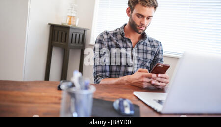 Incliné vers l'image d'un jeune entrepreneur de race blanche bien soigné à l'aide de son téléphone cellulaire occupé avec les deux mains tout en étant assis à son bureau derrière son ordinateur portable à son Banque D'Images
