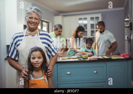 Grand-mère et petite-fille smiling at camera tandis que les membres de la famille preparing dessert en arrière-plan à la maison Banque D'Images