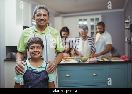 Grand-père et petit-fils smiling at camera tandis que les membres de la famille preparing dessert en arrière-plan à la maison Banque D'Images