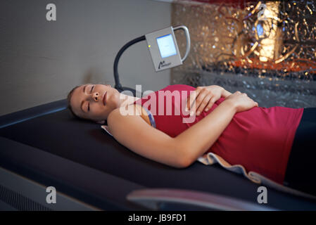 Woman in red shirt portant sur son dos sur table en salle de massage avec écran à côté de sa tête et de verre mur derrière, looking at camera Banque D'Images