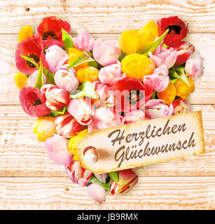 Souhaits d'anniversaire allemand sur un cœur floral coloré formé de tulipes au printemps frais dans des couleurs vives disposées sur un fond en bois rustique avec une étiquette disant - Joyeux Anniversaire ou Herzlichen Gluckwunsch Banque D'Images