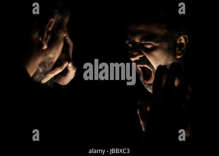 Homme en colère crier en face de miroir à son reflet dans une pièce sombre Banque D'Images