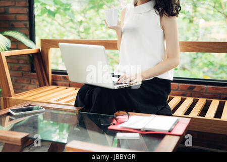 Confiant young asian woman dans smart casual wear shopping en ligne tout en s'asseyant à côté de fenêtre dans creative office ou cafe Banque D'Images