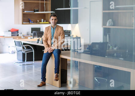 A 35 - 40 ans caucasien homme cheveux noirs cool look moderne en souriant un déshabillé chemise, jeans et blazer coin sur un bureau dans un bureau vide Banque D'Images