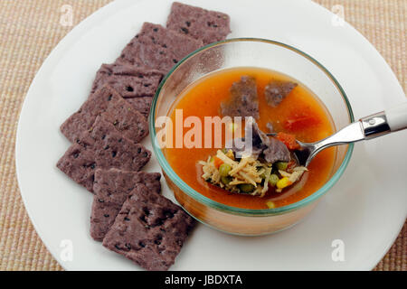 Spoon puiser dans un bol en verre de bouillon de poulet fait maison composé de poulet, pois, maïs, tomates, carottes et exotiques avec riz, riz violet toast, graines de sésame noir cracker carrés sur une plaque blanche. Banque D'Images