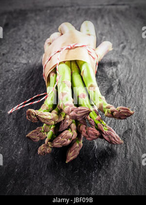 Asperges vertes fraîches livré dans du papier brun et liés avec de la ficelle affichés avec leurs conseils pour l'appareil photo sur une surface texturée gris ardoise Banque D'Images