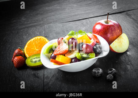 La préparation d'un délicieux bol de fruits frais colorés avec salade de fruits tropicaux, y compris les bleuets, pomme, orange, les kiwis et les fraises sur une surface ardoise cuisine Banque D'Images