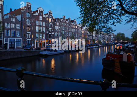 La photo montre un canal à Amsterdam dans la soirée avec quelques maisons anciennes et les brunchs d'un arbre. Les lumières de certaines lanternes reflète dans l'eau. Banque D'Images