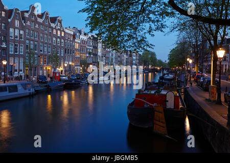 La photo montre un canal à Amsterdam dans la soirée avec quelques maisons anciennes et les brunchs d'un arbre. Les lumières de certaines lanternes reflète dans l'eau. Banque D'Images