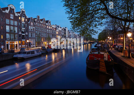 La photo montre un canal à Amsterdam dans la soirée avec quelques maisons anciennes et les brunchs d'un arbre. Les lumières de certaines lanternes reflète dans l'eau. Banque D'Images