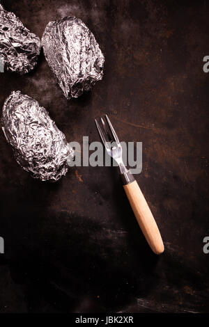 Enveloppé d'aluminium sur le barbecue encore des pommes de terre enveloppées dans leur papier d'aluminium d'argent portant sur une vieille ardoise surface avec une fourchette et copyspace, overhead view Banque D'Images