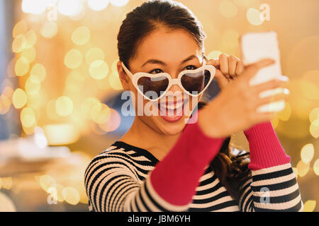 Chinois ludique femme portant des lunettes en forme de coeur en tenant avec selfies téléphone appareil photo Banque D'Images